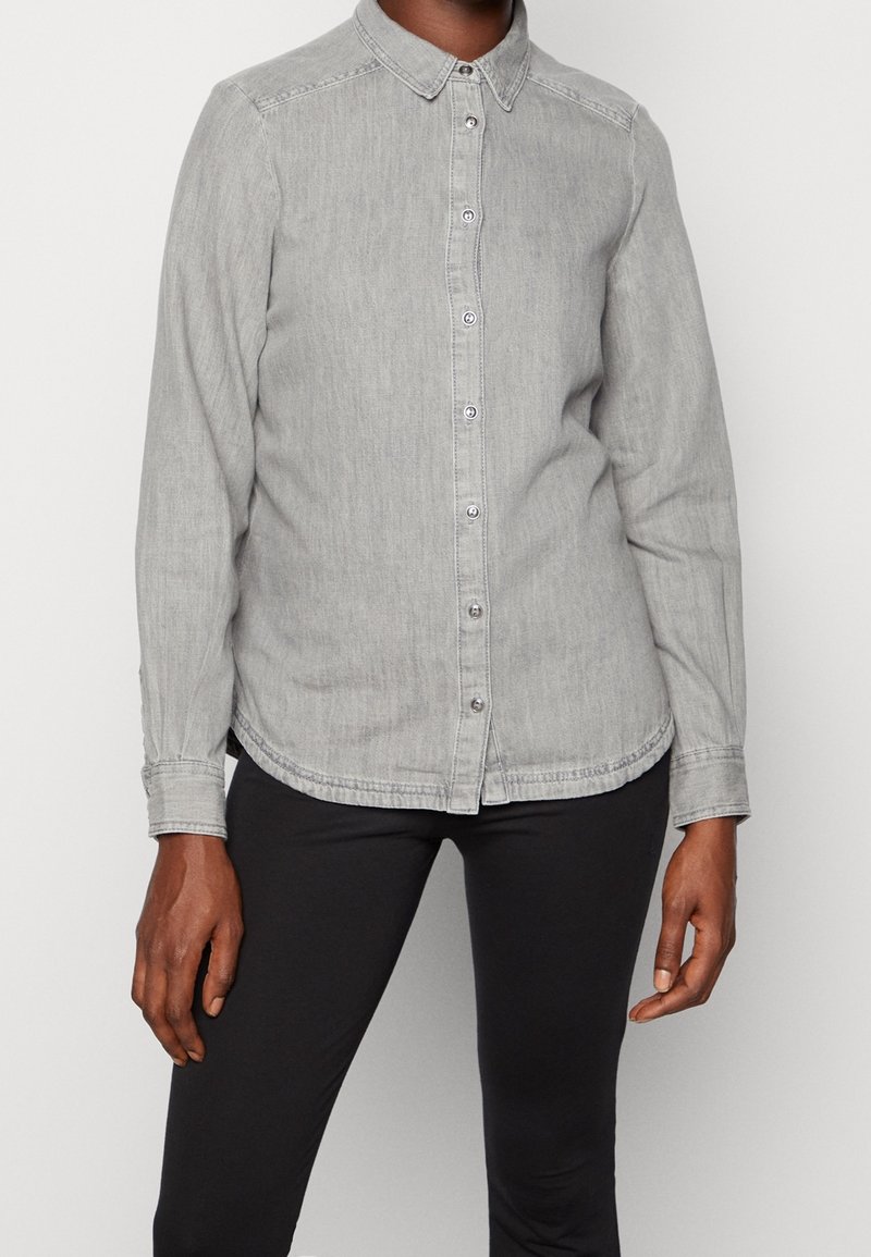 Grå denim skjorte med knapper, krave, lange ærmer, synlig syning og buet kant, parret med sorte bukser.