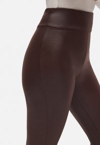 Leggings de tiro alto, de color marrón oscuro, con una textura lisa y brillante y un diseño ajustado. Presentan una cinturilla ancha y piernas entalladas.