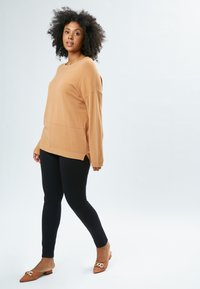 Lysebrun oversized sweater med lange ærmer, båret sammen med sorte leggings; lysbrune slip-on sko med en gylden detalje.