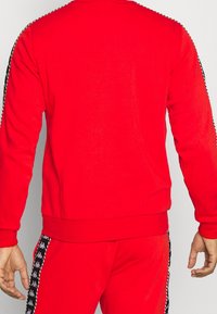 Sweatshirt rouge avec manches longues, ourlet côtelé et accents latéraux rayés noirs et blancs. Fabriqué en matière douce et texturée.