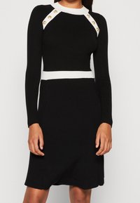 Vestido de punto acanalado negro con mangas largas, detalles en blanco en el escote y la cintura, que presenta botones dorados en los hombros.