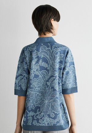 Persoon met kort donker haar die een blauw overhemd draagt met lichtblauwe ingewikkelde bloemmotieven, van achteren gezien tegen een effen achtergrond.