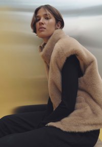 Beige Kunstpelz-Weste mit hohem Kragen, Reißverschlussdetails und seitlichen Schlitzen, getragen über einem schwarzen Langarmoberteil und dunklen Hosen. Glatter Hintergrund.