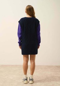 Granatowa dzianinowa sukienka z teksturowanym diamentowym wzorem, krótkie rękawy w kolorze fioletowym, długość powyżej kolan, noszona z szarymi sneakersami i białymi skarpetkami.