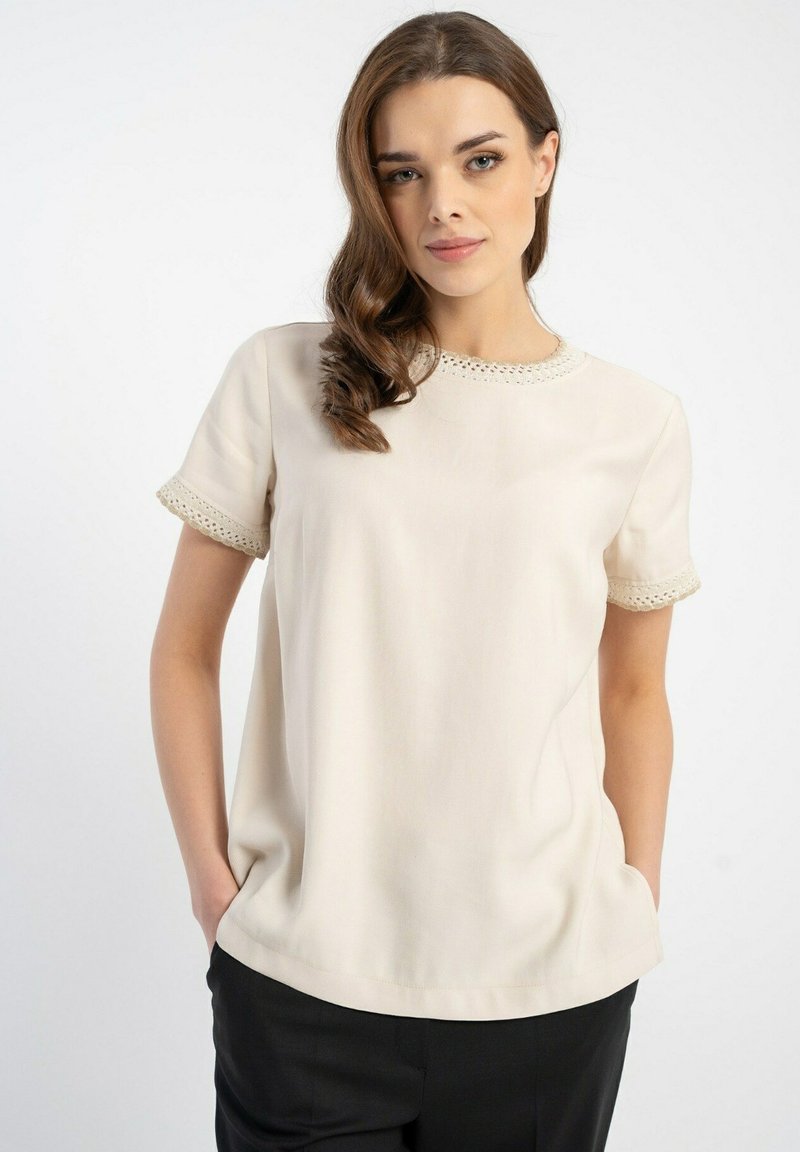 Beżowa bluzka z krótkim rękawem, koronkowymi wykończeniami na mankietach i dekolcie, z miękkiej tkaniny, luźny fason i delikatny połysk, w połączeniu z ciemnymi spodniami.