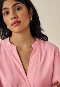 Blusa rosa con colletto, scollatura a V profonda, con una texture liscia, dettagli di cucitura sottili e maniche corte. Orecchini a cerchio semplici.