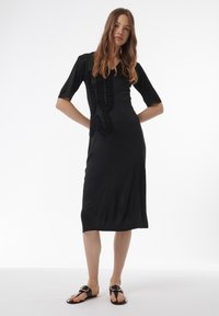 Zwart midi-jurk met korte mouwen, V-hals en een getextureerde ruffle-details aan de voorkant. Gecombineerd met zwarte platte sandalen.