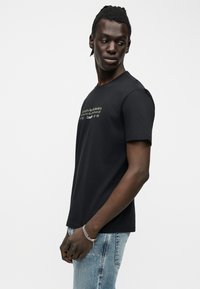 Homme aux cheveux tressés portant un T-shirt Calvin Klein noir avec un texte doré et un jean bleu clair, regardant par-dessus son épaule sur fond blanc.