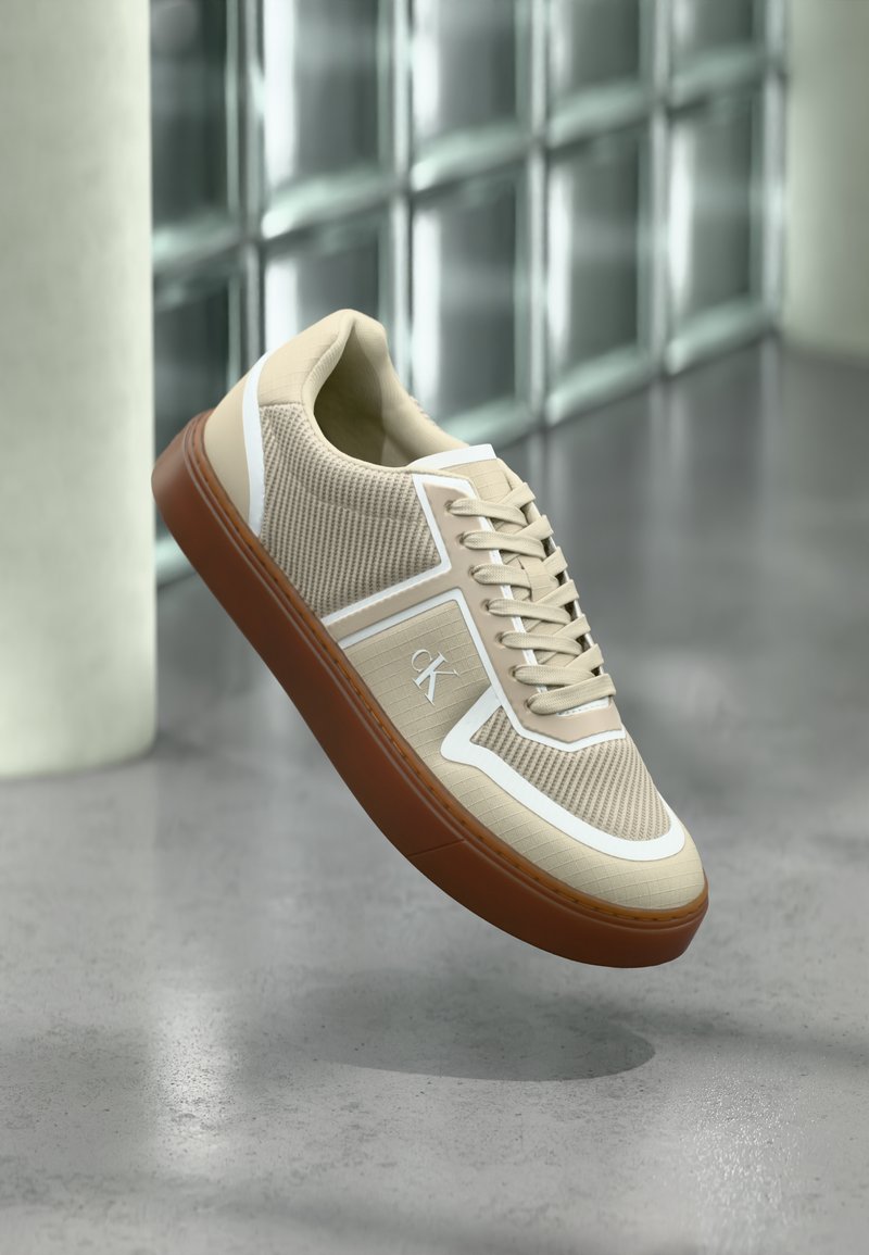 Μπεζ αθλητικό παπούτσι με λευκές λεπτομέρειες και καφέ σόλα αιωρούμενο πάνω από γυαλισμένο δάπεδο από σκυρόδεμα κοντά σε τοίχο από γυάλινα τουβλάκια σε ένα μοντέρνο εσωτερικό χώρο.