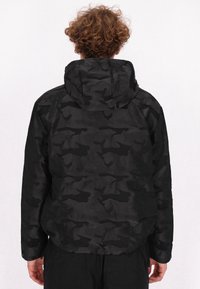 Schwarze camouflage Jacke mit Kapuze, aus strukturiertem Stoff und streamline Design. Die Rückansicht zeigt einfache Formen und Nähdetails.