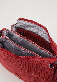 Sac bandoulière perforé rouge avec deux compartiments zippés et doublure en tissu gris de marque comportant un texte imprimé.