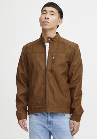 Veste en daim marron avec un col haut, fermeture éclair à l'avant et une poche poitrine. Présente une texture lisse et une silhouette ajustée.