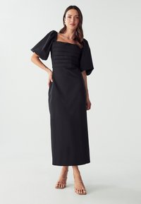 Black midi dress with a square neckline, structured pleated bodice, short puff sleeves, and a straight skirt, paired with nude sandals.