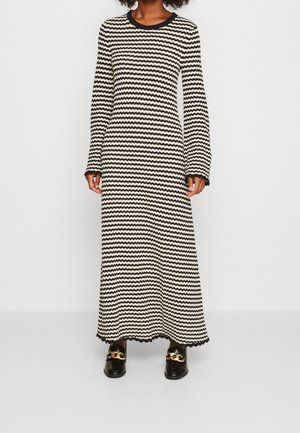 Frau trägt ein langes schwarz-weißes Kleid mit Zickzackmuster, langen Ärmeln und schwarzen Loafers mit goldenen Ketten.