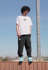 Witte t-shirt met korte mouwen en een klein zwart Nike-logo, gecombineerd met zwarte joggingbroek met een wit logo, en turquoise sneakers.