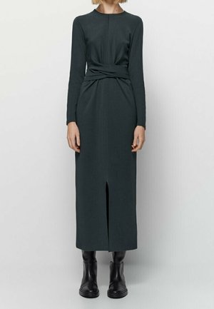 Robe midi à manches longues vert foncé pour femmes, avec fente avant et détail de taille torsadée, portée avec des bottines noires, présentée sur un modèle debout.