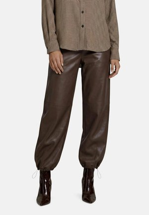 Personne portant un pantalon en cuir marron foncé avec chevilles froncées, associé à une chemise marron à carreaux boutonnée et des bottes marron foncé à talons.
