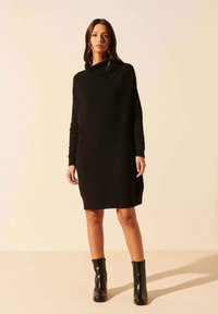 Robe tricotée noire à manches longues avec un col bénitier, présentant une coupe décontractée qui tombe au-dessus du genou. Associée à des bottines noires.