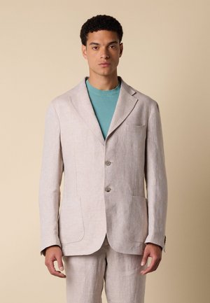 Jeune homme portant un costume en lin beige clair avec deux boutons et des poches plaquées sur un tee-shirt bleu sarcelle à col rond, debout devant un fond beige.