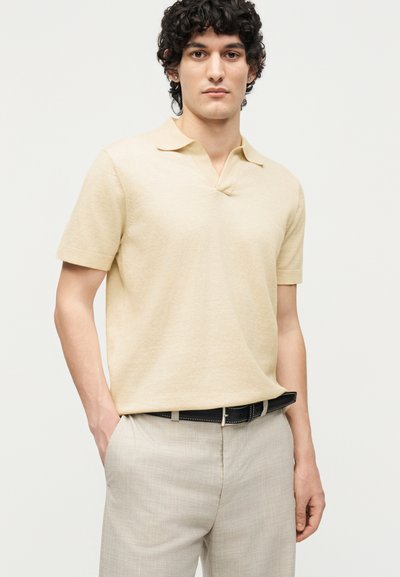 Νέος άνδρας με σγουρά σκούρα μαλλιά που φοράει ένα μπεζ κοντομάνικο πόλο πουκάμισο μπουκλωμένο σε ανοιχτόχρωμο γκρι παντελόνι με μαύρη ζώνη.