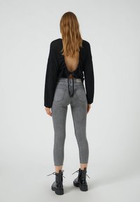 Suéter negro de espalda abierta con mangas largas y un detalle de lazo, combinado con jeans ajustados grises de talle alto y botas gruesas negras con cordones.