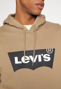 Personne portant un sweat à capuche beige Levi's avec un logo noir et blanc, montrant le cou et le bas du visage légèrement tournés sur le côté.