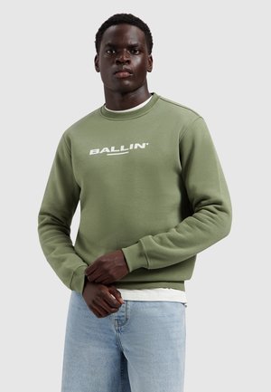 Man draagt een groen sweatshirt met de tekst "BALLIN'", lichtblauwe jeans, staand met samengevouwen handen tegen een grijze achtergrond.