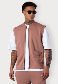 Hellrosa ärmellose Jacke aus weichem Stoff mit einem Reißverschluss vorne, zwei Fronttaschen und gesticktem Logo. Getragen über einem weißen T-Shirt.