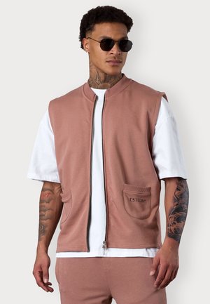 Hellrosa ärmellose Jacke aus weichem Stoff mit einem Reißverschluss vorne, zwei Fronttaschen und gesticktem Logo. Getragen über einem weißen T-Shirt.