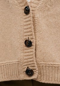 Cárdigan de punto beige con siete grandes botones negros, bordes acanalados y un acabado texturizado. Detalle de diseño cosido a lo largo de la tapeta de botones.