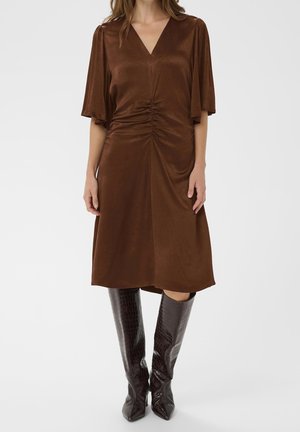 Femme portant une robe midi marron à col en V avec un devant froncé et de larges manches évasées, associée à des bottes mi-hautes marron foncé texturées.