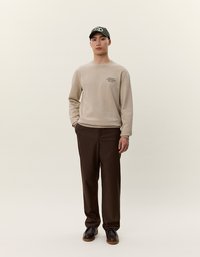 Sudadera beige con logo, pantalones marrones y gorra verde. El conjunto presenta un ajuste relajado, textura mínima y elementos de diseño clásicos.