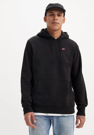 Sudadera negra con cordones y bolsillo frontal, con un pequeño logo rojo. Combinada con jeans de lavado claro. Textura suave, ajuste casual.