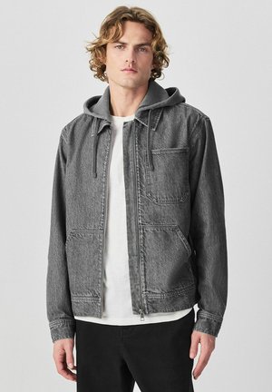 Mladý muž v sivej rifľovej bunde s kapucňou, bielom tričku a čiernych nohaviciach stojaci pred jednoduchým svetlým pozadím.