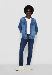 Chaqueta de mezclilla con dos bolsillos en el pecho, llevada sobre una camiseta blanca, combinada con jeans de color azul oscuro y zapatillas de color claro; unas gafas de sol negras completan el look.