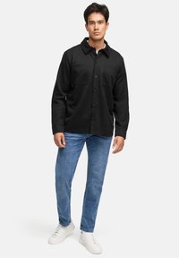 Svart knapp-up jakke med to brystlommer, båret over en hvit skjorte, kombinert med blå jeans og hvite sneakers. Glatt tekstur.