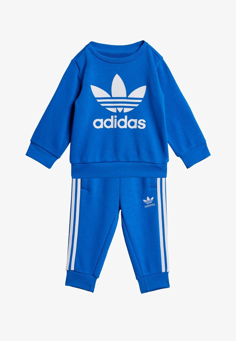 Compleu sportiv albastru format dintr-un pulover cu guler rotund și pantaloni subțiri. Puloverul are un logo alb mare; pantalonii au dungi albe pe margine.