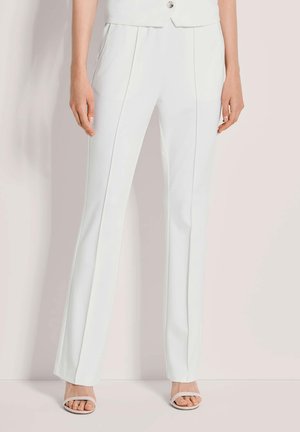 Weiße, taillierte Damenhose mit Bügelfalten vorne, kombiniert mit weißen offenen Sandaletten vor hellem Hintergrund.