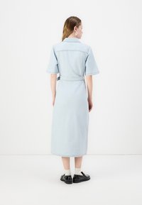 Lyseblå denimkjole med korte ermer, belte i midjen og lang, rett fasong. Svarte sko med synlige hvite sokker ved kanten.