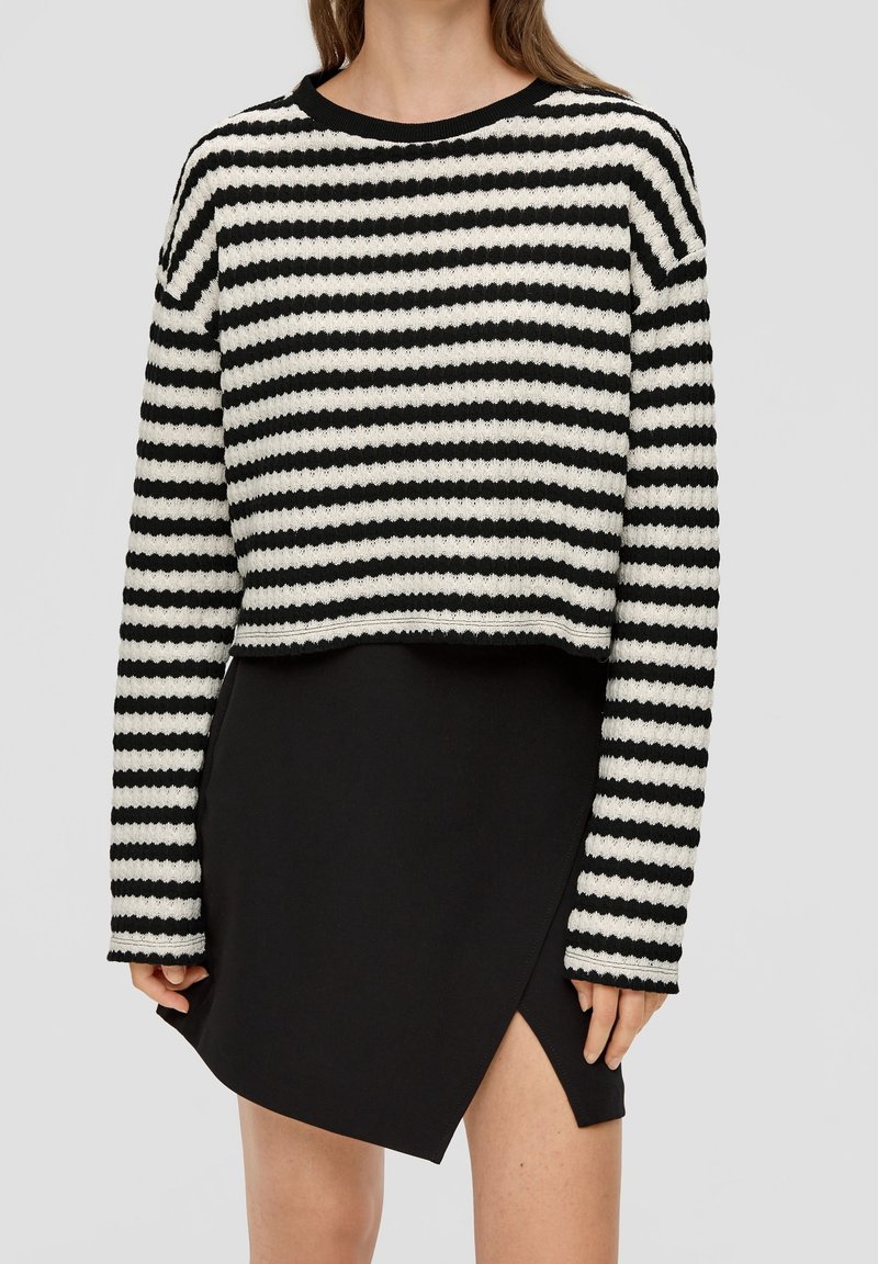 Pull rayé noir et crème avec un tricot texturé, associé à une jupe noire fendue sur le côté. Le pull est court et possède des manches longues.