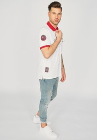 Vit polo-skjorta med röd krage och muddar, med ett märke på ena ärmen och en etikett vid fållen. Bärs med ljusblå slitna jeans och vita sneakers.