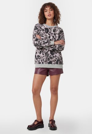 Kvinde med krøllet hår iført stjerneformede øreringe, leopardprint trøje, bordeaux læder shorts og chunky bordeaux sko, stående med armene krydset.