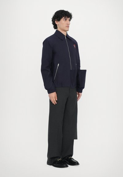 Giacca zip-up blu navy con colletto, polsini elasticizzati e una tasca laterale. Presenta un emblema ricamato sul petto, realizzata in materiale leggero.