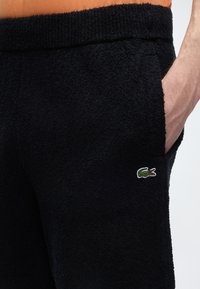 Gros plan d'une personne portant un short noir texturé avec un petit logo crocodile vert et une main dans la poche.