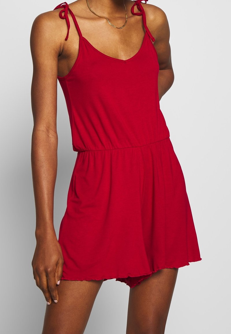 Femme portant une combinaison rouge sans manches avec des bretelles nouées aux épaules et une taille élastique, debout devant un fond clair uni.
