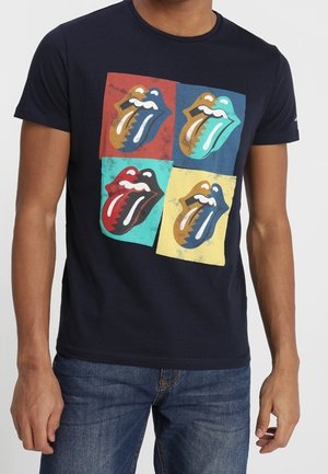 Hombre con camiseta azul marino con cuatro logotipos coloridos de la lengua de los Rolling Stones en cuadrados rojo, azul, amarillo y verde azulado en el pecho.