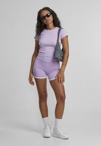 T-shirt aderente viola con logo bianco, pantaloni corti a vita alta abbinati, rifiniture bianche e sneakers bianche. Occhiali da sole neri e borsa grigia.
