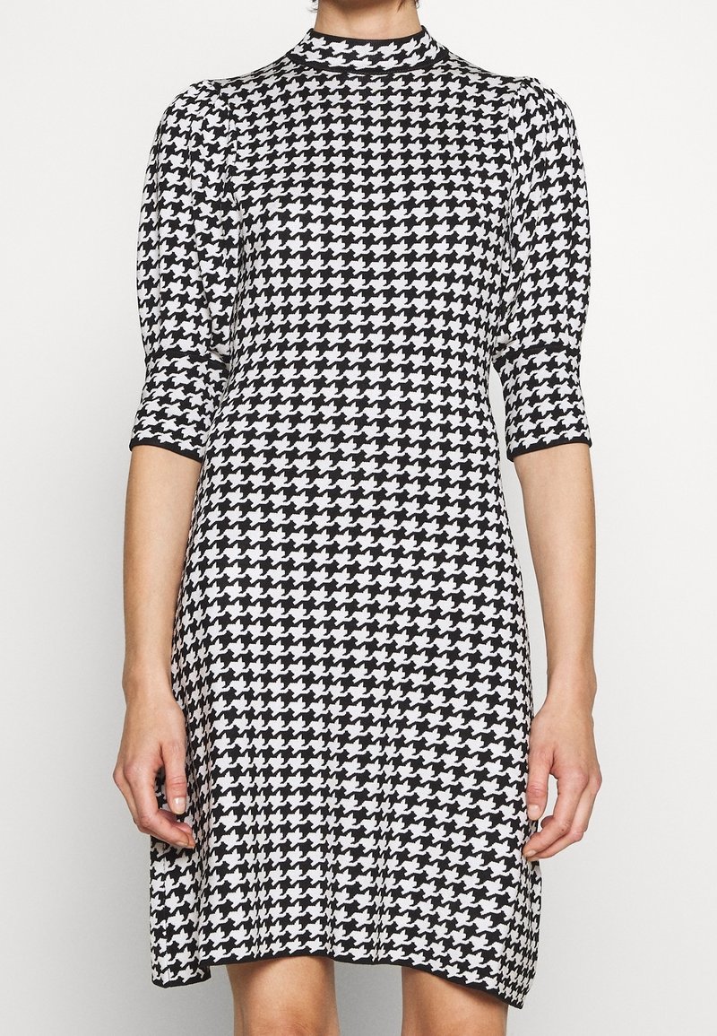 Femme portant une robe noire et blanche à motif pied-de-poule, longueur genou, manches trois-quarts et col montant.