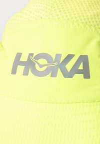 Φωτεινό κίτρινο ύφασμα με γκρι ανακλαστικό λογότυπο HOKA και σχέδιο με στυλιζαρισμένο πουλί, ορατά ράμματα και υφή διχτυού πάνω από το λογότυπο.