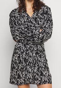 Svart omlottklänning med vitt abstrakt mönster, långa ärmar, V-ringning och gathered midjedetalj. Mjuk tyg med en flytande passform.